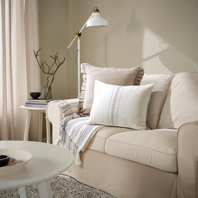 Cosy living room with beige sofa, ÅKERNEJLIKA cushion, decorative pillows, side table, and floor lamp.
