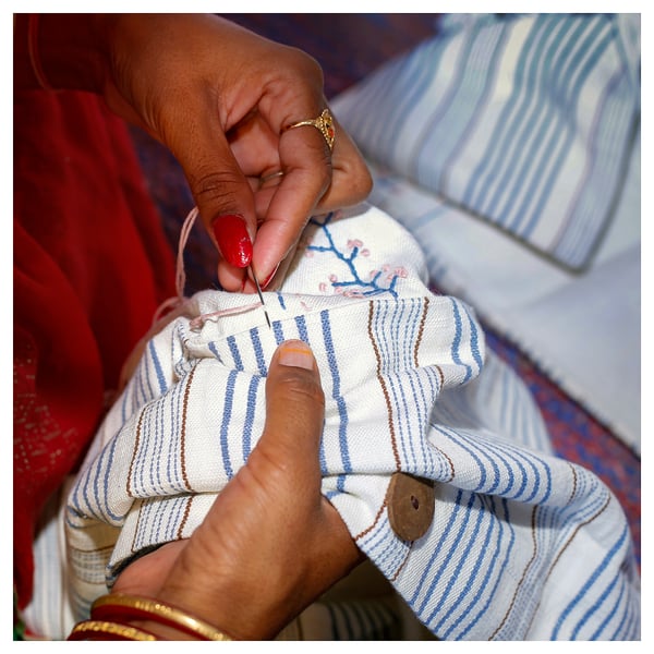 Hands sew intricate floral pattern on a white, blue-striped cushion cover. Red nails, gold ring, and bracelets are visible.
