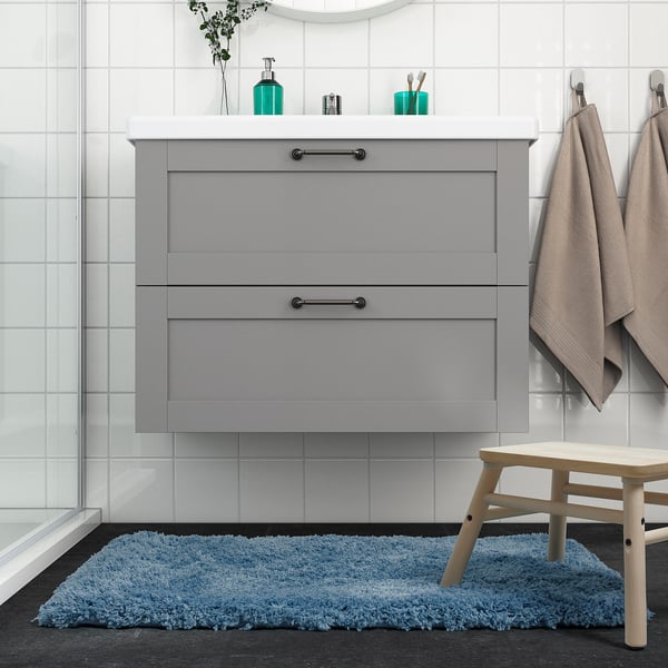 Modern bathroom with ALMTJÄRN blue bath mat. Grey vanity, white tiles, wooden stool, beige towels.