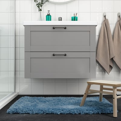 Modern bathroom with ALMTJÄRN blue bath mat. Grey vanity, white tiles, wooden stool, beige towels.