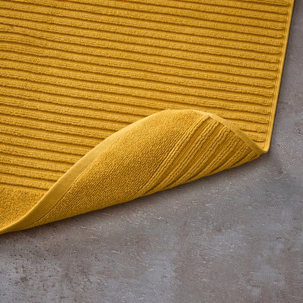 Mustard yellow bath mat made of pure cotton terry. Its structured weave massages feet, absorbs well, and dries quickly.