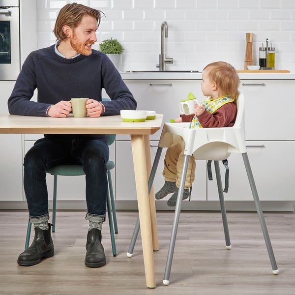 Person in blue sweater sitting at table with coffee, next to toddler in highchair eating from green bowl.