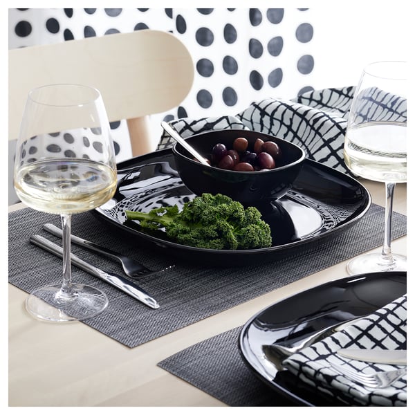 Monochrome table setting with BACKIG black plates, bowls, wine glasses, and cutlery on dark mats.