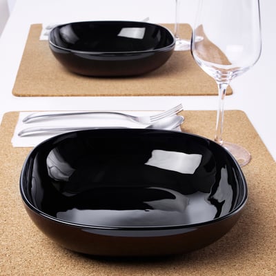 Two black glossy bowls on cork placemats with utensils and wine glasses, suitable for setting a stylish table.