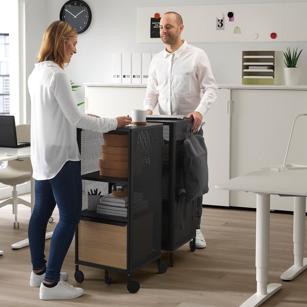 Two people in an office discuss documents at a black BEKANT mobile cart with wheels, storing files and a mug, near a standing desk.