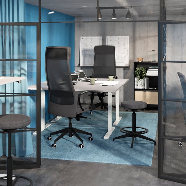 Office meeting room with white BEKANT table, grey chairs, blue rug, and concrete walls. Glass partitions with black frames.