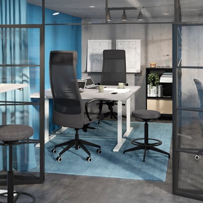 Small office. Glass walls, blue curtains. Workspace with white BEKANT table, grey chairs, laptop, cups. Blue rug. Plant shelf.