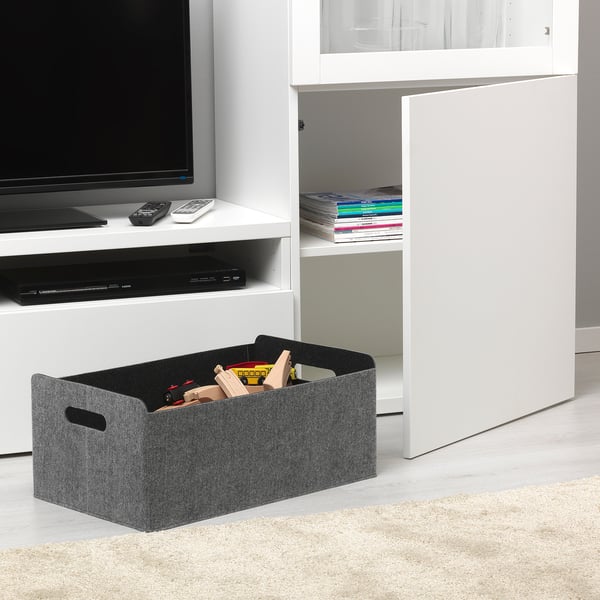 Living room scene with a grey felt storage box full of toys beside a white tv stand holding a flat-screen tv, electronics, and magazines.
