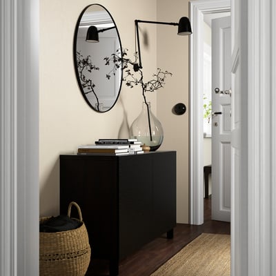 Modern entryway with large black BESTÅ sideboard; woven basket, black wall mirror, black wall sconce, glass vase with branches.