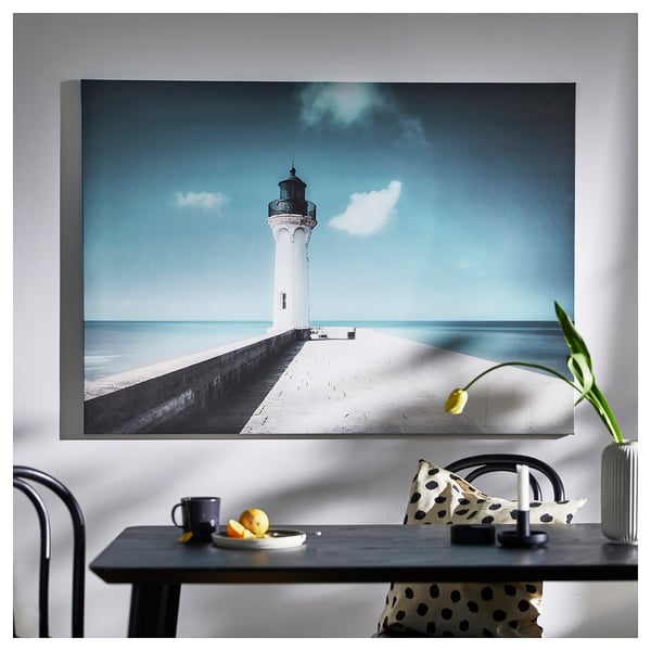 Large framed canvas a lighthouse near ocean on wall above table with a mug and fruits.
