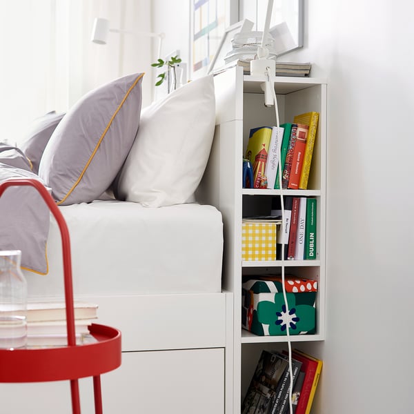 Bedroom scene with BRIMNES white bed, purple pillows, red side table, books, and plants.