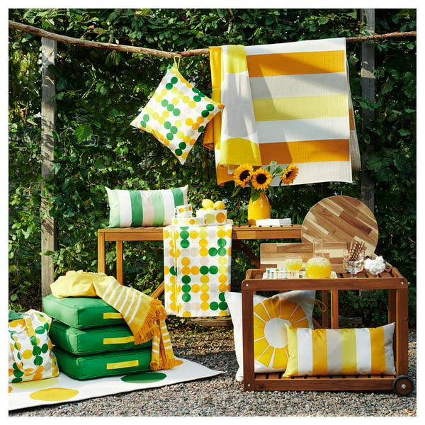 Bright yellow and green outdoor decor setup with BRÖGGAN recycled polyester cushions, blankets, and a wooden cart on gravel, surrounded by greenery.