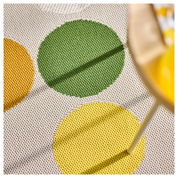 Close-up of a colourful, textured rug with green and yellow dots on a beige grid. A hand holds a remote near it.