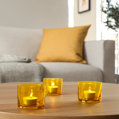 Three yellow glass candle holders on a wooden table, each containing a lit tea light, creating a warm and cosy ambiance.