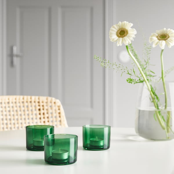 Three green glass candle holders with small candles on white table beside white vase with daisies.
