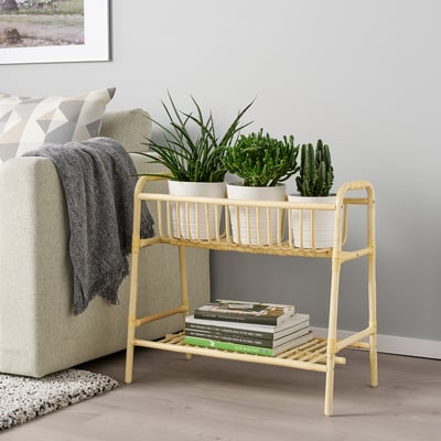Rattan plant stand in beige with books and plants near a sofa.