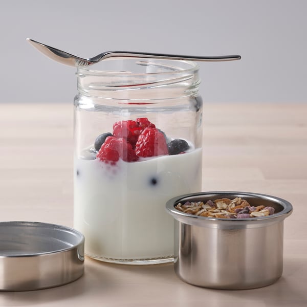 Glass jar with fruit yoghurt and berries, accompanied by metal containers for dry foods. Spoon on jar lid, ideal for breakfast on-the-go.