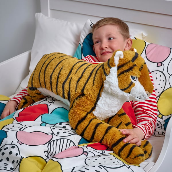 A boy lies on a bed hugging a large stuffed tiger. The tiger is designed for comfort and play, perfect for children aged 18 months and up.