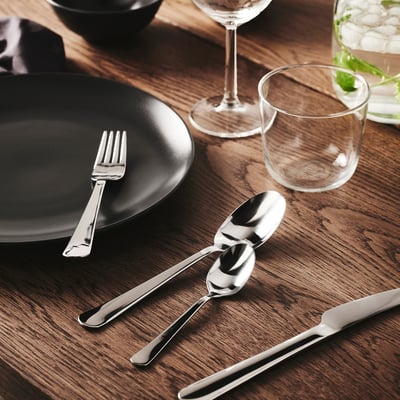 Table setting with shiny metallic fork, two spoons, and knife on dark wooden table, accompanied by black plate and glass drinkware.