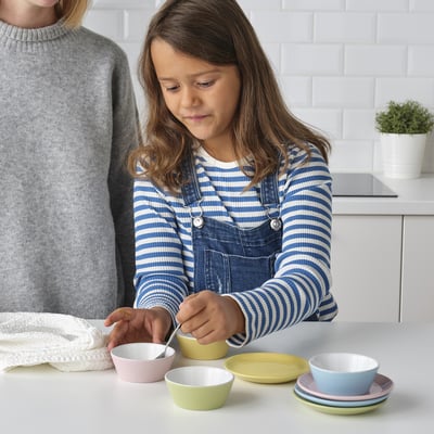 Child in blue coveralls plays with colourful bowls using a spoon on kitchen counter, promoting role-play and fine motor skills.