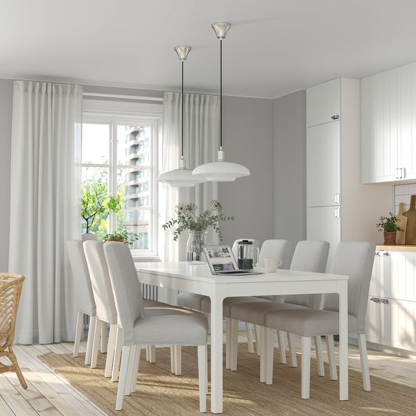 Modern white dining set with extendable table and grey chairs, large window with curtains, white pendant lights, potted plants, and kitchen cabinets.