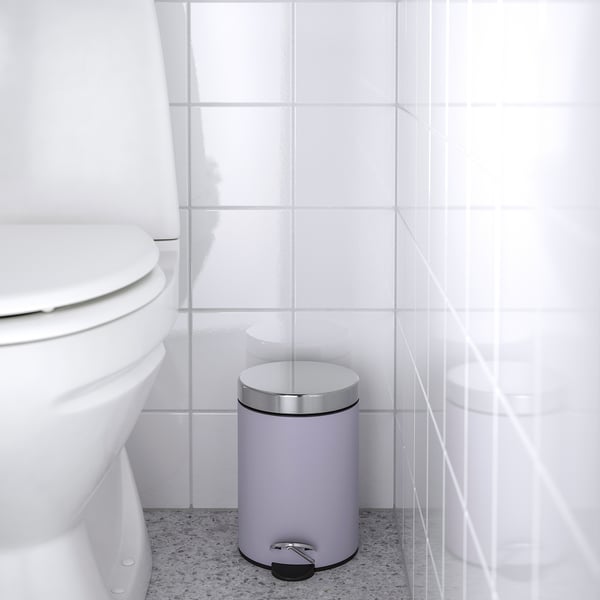 A white bathroom with a closed toilet seat and a grey rubbish can with a foot pedal and silver lid next to it.