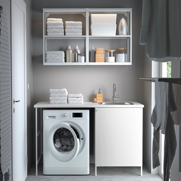 Modern laundry set-up. White ENHET unit with sink, washing machine, shelves, and storage. Grey curtains, folded towels, and clean design.
