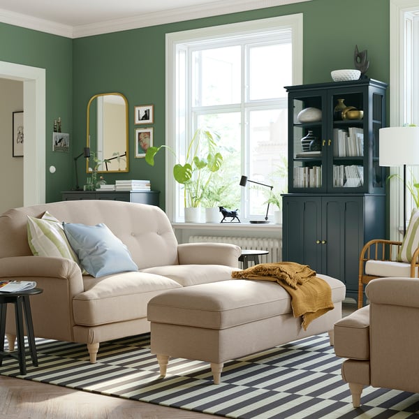 Cosy living room with green walls, beige sofa, striped rug, dark wooden cabinet, and large window.