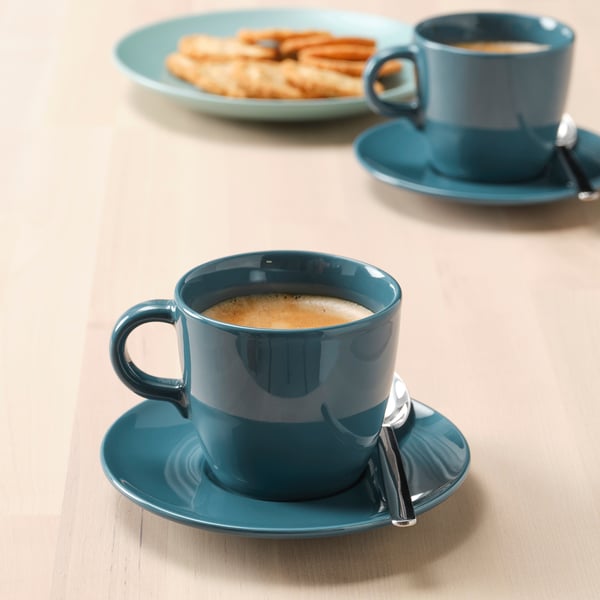 Two teal ceramic cups and saucers with coffee, one with a spoon, and a plate with pastries on a light wooden table.