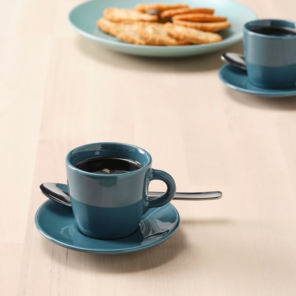 Two teal cups with saucers, one with a spoon, likely filled with coffee or tea. A plate with pastries in the background.
