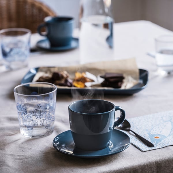 Table setup with FÄRGKLAR cups in blue, one filled with steaming coffee, matched with clear patterned glasses, and a tray of food on the side.