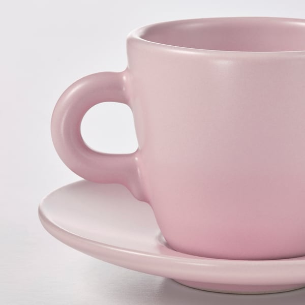 A close-up of a single, simple, pastel pink cup with a matching saucer, showcasing smooth edges and a glossy finish. Positioned on a white surface.