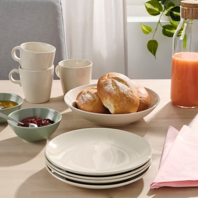 Wooden table set with FÄRGKLAR side plates, bowls, mugs, bread, jam, and juice.