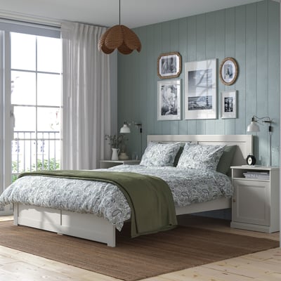Bedroom set: white bed, green blanket, wooden table, lamp, art, window curtains.