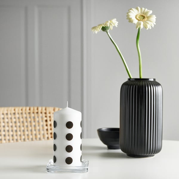 A FENOMEN white polka dot candle in clear stand sits near a tall black ribbed vase with two white flowers and a small black bowl.
