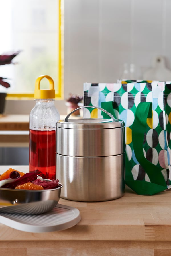 Image with stainless steel FÖRSKAFFA tiffin box, colourful dish, and drink bottle on a wooden table.