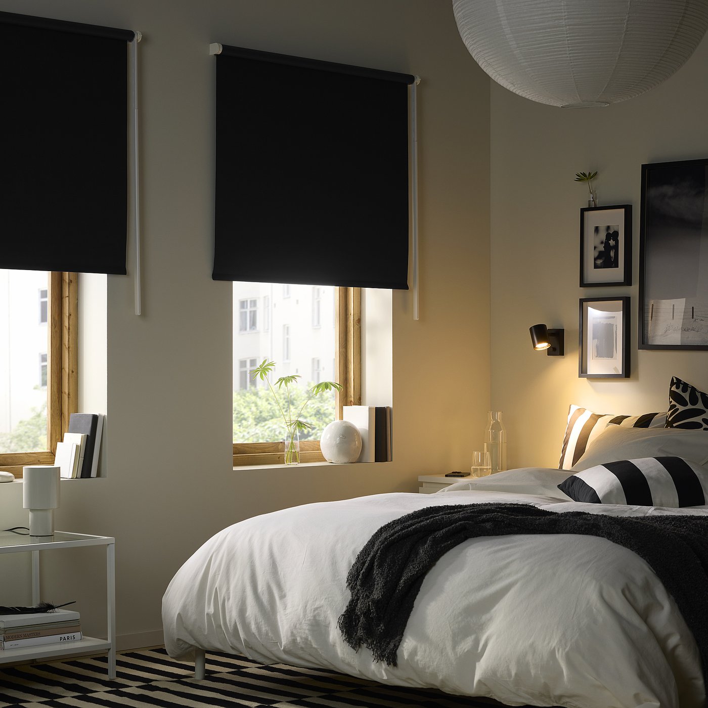 Bedroom with 2 black roller blinds on large windows, bed with white linens, and a cosy bedside setup.
