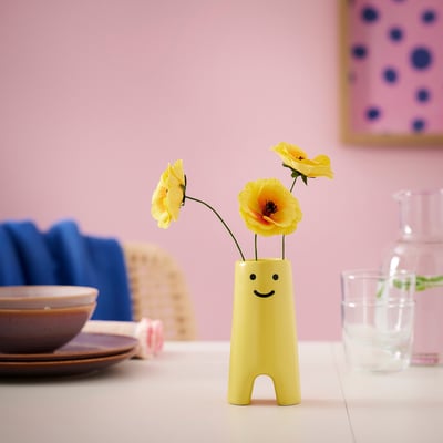 Small, yellow, stoneware vase named FRÖVÄXT, with a face design, holding yellow flowers, sitting on a table with plates and a glass.