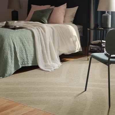 Bedroom with light beige FULLMAKT rug, white bed, green pillows, dark grey chair. Rug is large, covering wooden floor.