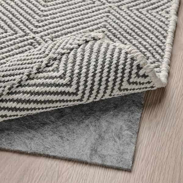 A folded grey and white woven rug, displayed on a wooden floor, with a felt underlay visible.