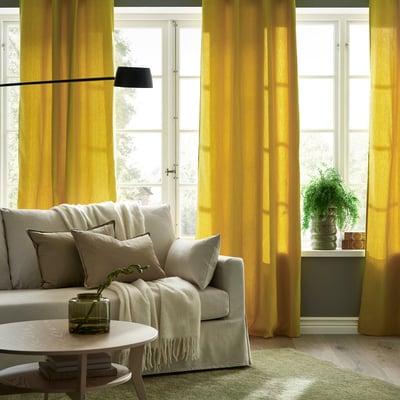 Yellow curtains frame windows; grey sofa with beige pillows, white blanket; small plant adds greenery.