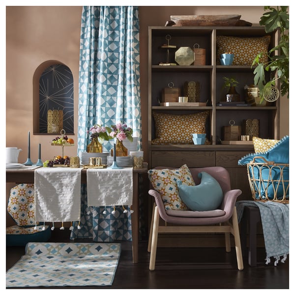 Bright living room with blue & yellow patterns, includes comfortable chair, colourful pillows, bookshelf with decorations, plants, and a vibrant rug.