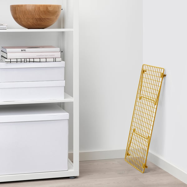 Yellow GREJIG shoe rack, leaning against white wall, displayed next to organised shelves with books and boxes.