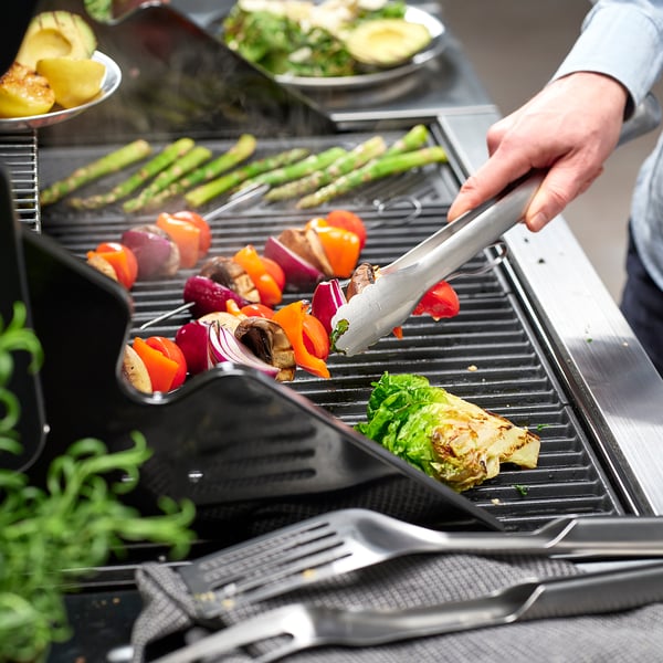 Hand grilling colourful veggies with tongs on a modern grill next to other cooking tools.