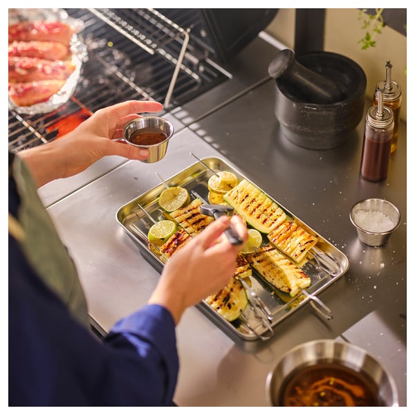 Person seasoning grilled skewers on stainless steel tray next to grill, with small bowls of spices and sauce nearby.