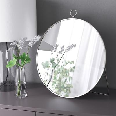 Round mirror stands on counter, reflecting lamp and flowers. Has hanging ring on top.