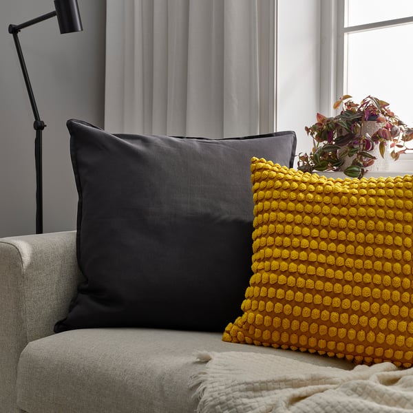 Two throw pillows on a sofa: black square & large yellow textured square, with a black standing lamp & potted plant nearby.