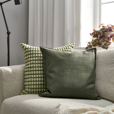 A cosy living room with an olive green GURLI cushion on a light sofa, enhancing decor with textured design and soft fabric.