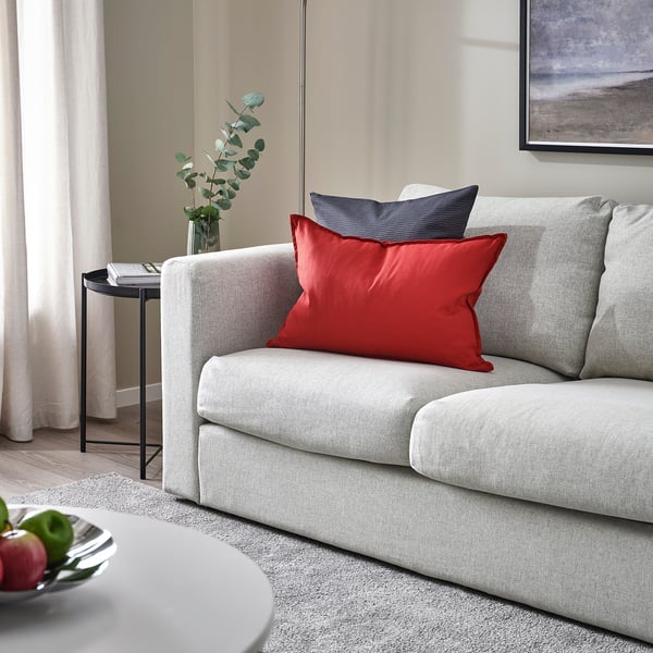Modern living room with grey GURLI sofa, red/blue cushion, side table, green plant, rug, wall art, white curtain.