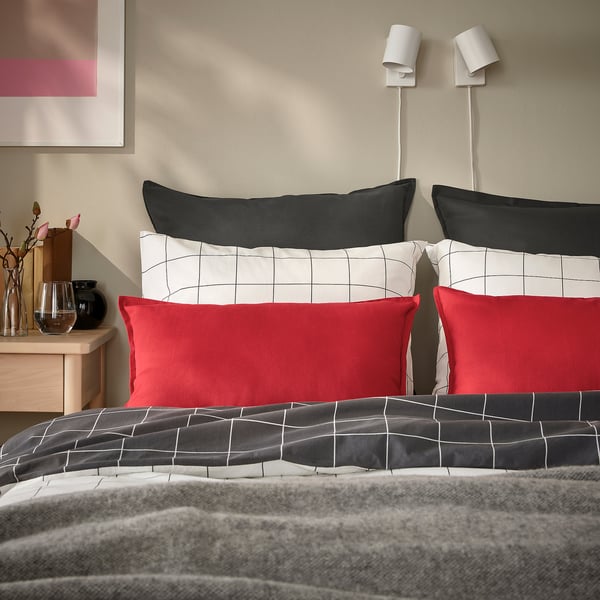 Cosy bed with red & grid pillows, grey fur blanket. Two wall lamps, art. Side table with vases & flowers.